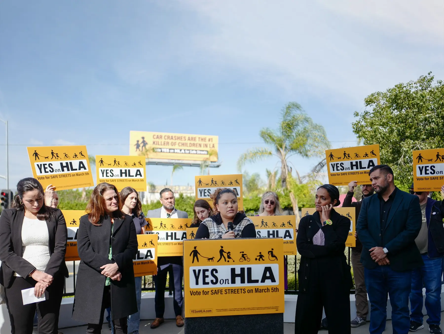 A group of HLA Activists, electeds, and coalition partners stand before a podium holding bright yellow 'Yes on HLA' signs. Behind them are palm trees, a blue sky, and a yellow billboard that says "Car Crashes are the #1 Killer of Kids in LA". 